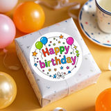 Gift box with a 'Happy Birthday' sticker surrounded by balloons on a yellow surface